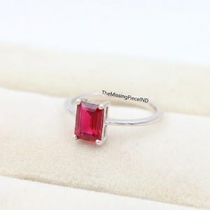 May include: A silver ring with a rectangular, bright red gemstone. The gemstone is the main feature, set in a simple, elegant band. The ring is shown on a soft, off-white surface.