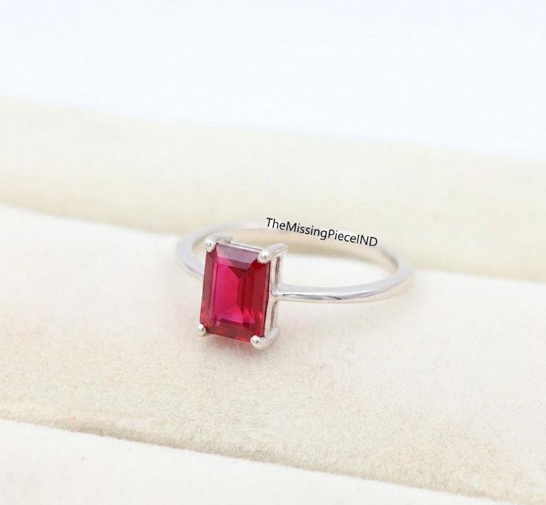 May include: A silver ring with a rectangular, bright red gemstone. The gemstone is the main feature, set in a simple, elegant band. The ring is shown on a soft, off-white surface.