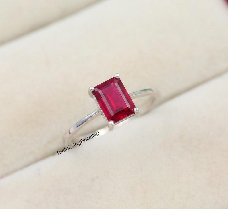 May include: A silver ring with a rectangular, deep red gemstone. The gemstone is the main feature, set in a simple, elegant band. The ring is shown on a soft, light-coloured surface, emphasising its design.