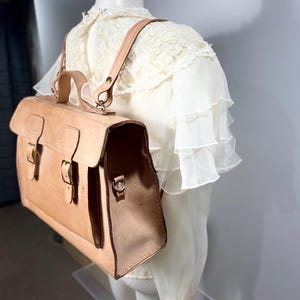 May include: A light brown leather satchel with a top handle and adjustable shoulder straps. The bag features two front buckles and a rectangular shape. The bag is displayed on a mannequin wearing a white blouse with lace and ruffled sleeves.