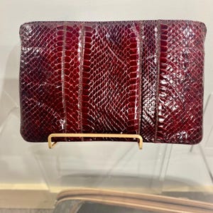 May include: A rectangular, burgundy clutch with a snakeskin pattern. The clutch is displayed on a gold-colored stand. The snakeskin pattern is a deep red with a glossy finish, and the clutch appears to be made of leather.