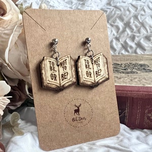 May include: A pair of wooden book earrings with the text "To be, or not to be?" on each earring. The earrings are on a brown card with the text "Oh Deer" in a circle.