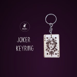 May include: Wooden keyring featuring a Joker playing card design with a detailed illustration of the Joker. The keyring has a silver metal ring.