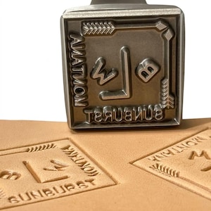 May include: A square metal leather stamping tool with a raised design featuring the letters B and W, the word MONTANA, and the word SUNBURST. The stamp is positioned above two identical impressions pressed into a piece of light tan leather.