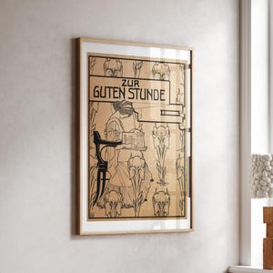 May include: A framed art print featuring a black line drawing on a tan background. The illustration depicts a person sitting in a chair reading, surrounded by stylized floral motifs. The text "ZUR GUTEN STUNDE" is printed in black at the top.