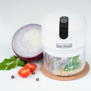 May include: A white electric food chopper with a clear plastic bowl and a black power button on top. The device features the brand name sarmali printed on the front. The chopper contains chopped ingredients and is placed on a round cork coaster.