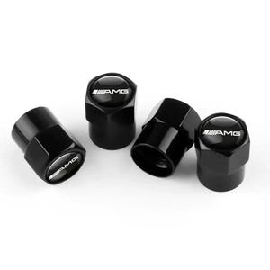 May include: A set of four black hexagonal tire valve caps with a glossy finish. Each cap features a circular top insert displaying the white AMG logo. The caps have a threaded interior design for secure attachment to vehicle tire valves.