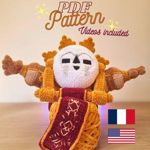 Peut inclure: Une poupée au crochet faite à la main avec un visage blanc, une crinière orange et des bras bruns. La poupée a une écharpe rouge et dorée. Le texte "PDF Pattern Videos included" est en haut de l'image.