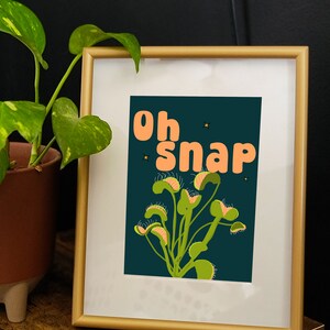 May include: A framed print with a dark green background and the text "Oh Snap" in orange letters. The print features a stylized illustration of a Venus flytrap plant in green and yellow.