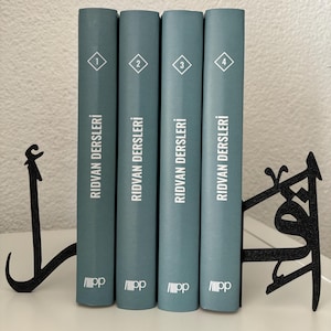 Könnte beinhalten: Vier blaue gebundene Bücher mit weißem Text und Zahlen auf den Rücken. Die Bücher sind mit "Ridvan Dersleri" betitelt und von 1 bis 4 nummeriert. Die Bücher werden von zwei schwarzen Metallbuchstützen in Ankerform gehalten.