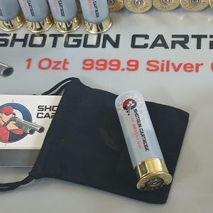 Puede incluir: Cartucho de escopeta plateado con detalles dorados, presentado con una bolsa negra y una tarjeta con un tirador. El texto "SHOTGUN CARTRIDGE" y "1 Ozt 999.9 Silver Gra" son visibles.