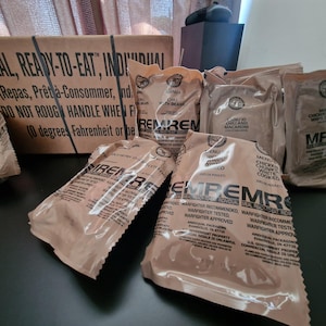 May include: A selection of brown MRE (Meal, Ready-to-Eat) food pouches and a cardboard box. The box has text in English and French. The pouches are labelled with menu details and the text "EMPEMR".