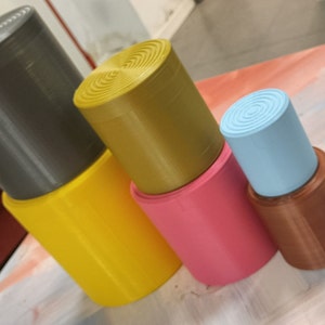 Op de afbeelding: Een set van vijf gestapelde cilindrische containers in verschillende kleuren, waaronder grijs, geel, roze, bruin en blauw. De containers zijn gemaakt van een glad, getextureerd materiaal.