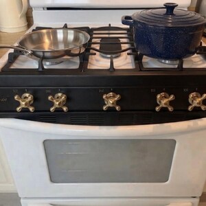 Puede incluir: Una cocina de gas blanca con panel de control negro y cinco mandos en forma de cruz color latón. Una sartén de acero inoxidable descansa sobre el quemador izquierdo y una olla de hierro fundido esmaltado azul oscuro moteado está a la derecha.