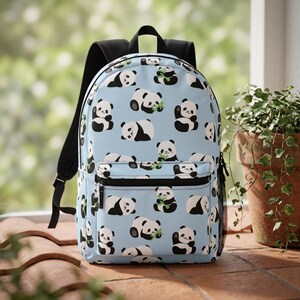 May include: A light blue backpack featuring an all-over pattern of cartoon pandas in various poses, some eating green bamboo stalks. The bag includes a front zipped pocket, black shoulder straps, and black mesh side pockets for storage.