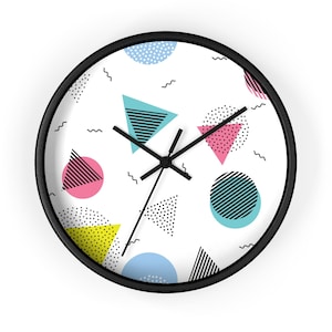 May include: A round wall clock with a black frame. The clock face is white and features colorful geometric shapes in pink, blue, yellow, and black. The clock hands are black.