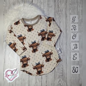 Könnte beinhalten: Ein beigefarbenes Langarmkleid mit Rundhalsausschnitt und einem Muster aus braunen Highland-Kühen mit blauen Schleifen auf weißem Polka-Dot-Hintergrund. Das Kleid hat einen leicht ausgestellten Rock und ist mit "Größe 80" gekennzeichnet.