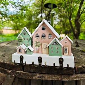 Op de afbeelding: Een decoratieve sleutelhouder met een verzameling miniatuurhuisjes in pastelkleuren. De huisjes zijn gemonteerd op een witte basis met bronzen haken. De achtergrond is een vage buitenscène met groen.