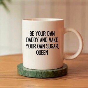 May include: A white ceramic mug with a handle, featuring the black printed text: BE YOUR OWN DADDY AND MAKE YOUR OWN SUGAR, QUEEN. The mug is cylindrical with a smooth finish and sits upright, displaying the bold, sans-serif lettering clearly.