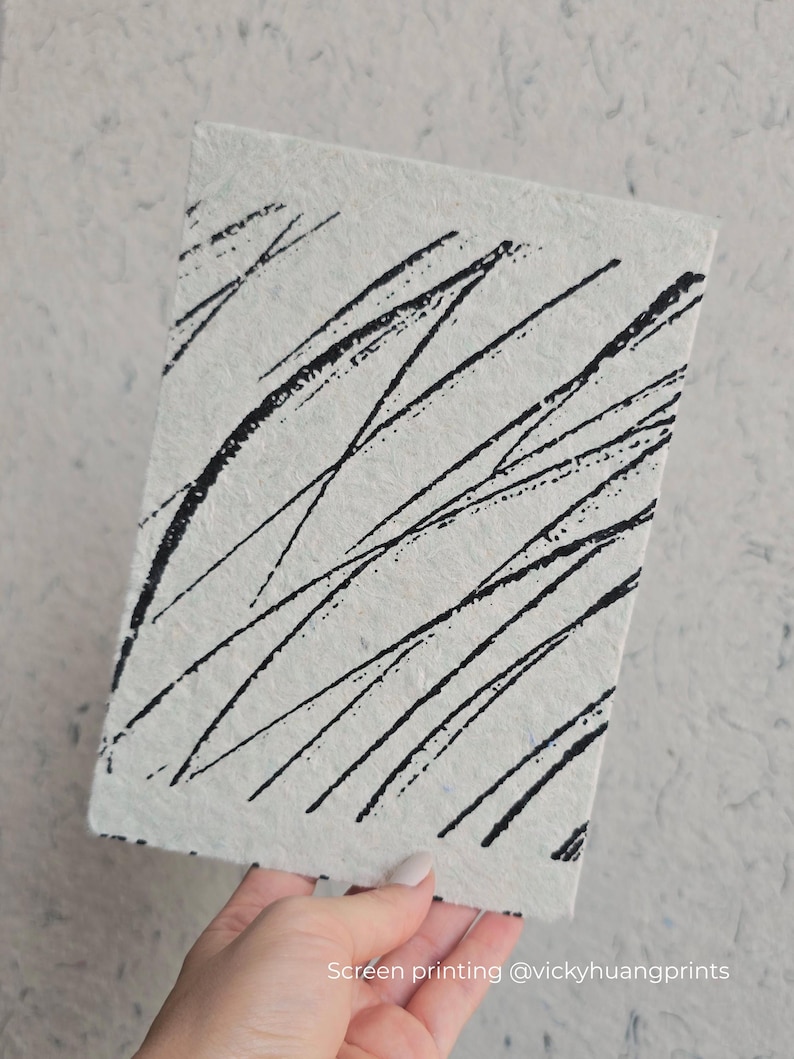 K&ouml;nnte beinhalten: Ein rechteckiges, cremefarbenes Notizbuch mit strukturierter Oberfl&auml;che. Auf dem Cover sind kr&auml;ftige, schwarze diagonale Linien aufgedruckt. Der Text "Screen printing @vickyhuangprints" ist unten rechts aufgedruckt.