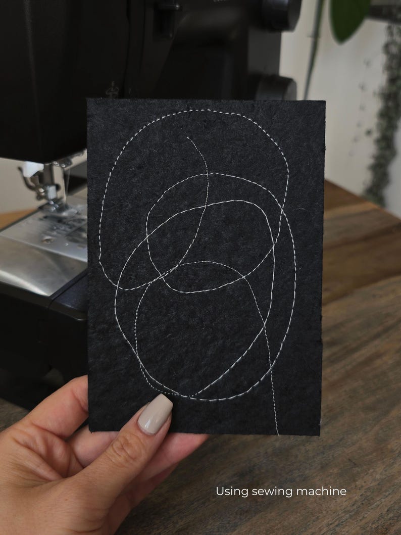 K&ouml;nnte beinhalten: Ein schwarzes, rechteckiges St&uuml;ck Stoff mit wei&szlig;en F&auml;den, die ein abstraktes Muster bilden. Der Text "Using sewing machine" ist unten sichtbar. Im Hintergrund ist eine N&auml;hmaschine zu sehen.