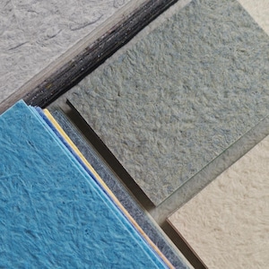 K&ouml;nnte beinhalten: Eine Sammlung von strukturierten Papierproben in verschiedenen Farben, darunter Blau-, Grau- und Off-White-T&ouml;ne. Das Papier hat ein faseriges Aussehen, was auf ein handgefertigtes oder recyceltes Material hindeutet. Die Proben sind in Stapeln und einzelnen Bl&auml;ttern angeordnet.