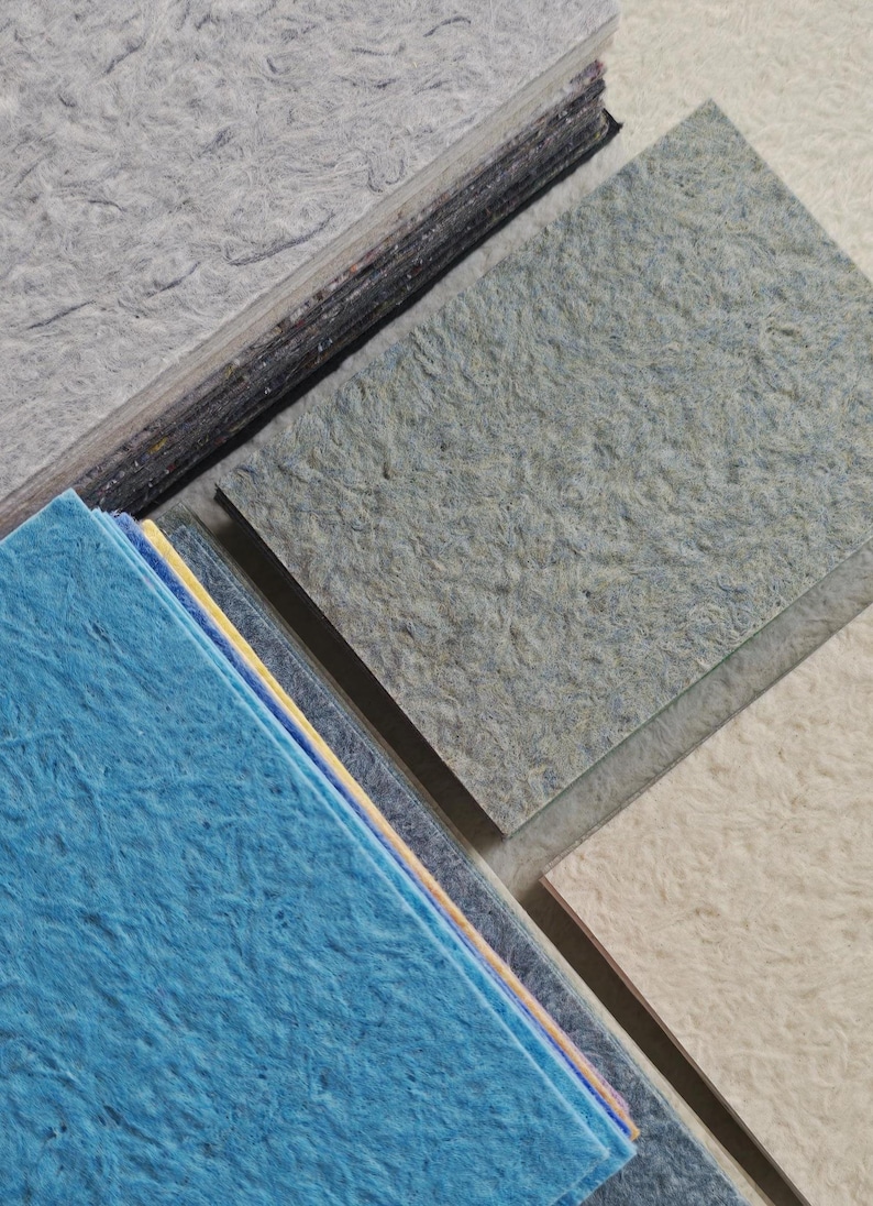 K&ouml;nnte beinhalten: Eine Sammlung von strukturierten Papierproben in verschiedenen Farben, darunter Blau-, Grau- und Off-White-T&ouml;ne. Das Papier hat ein faseriges Aussehen, was auf ein handgefertigtes oder recyceltes Material hindeutet. Die Proben sind in Stapeln und einzelnen Bl&auml;ttern angeordnet.