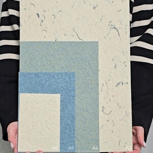 K&ouml;nnte beinhalten: Eine Sammlung strukturierter Papierb&ouml;gen in verschiedenen Gr&ouml;&szlig;en, darunter A3, A4, A5 und A6. Die Papiere weisen ein gesprenkeltes, organisches Design mit einer Mischung aus hellen Beige- und Blaut&ouml;nen auf. Die Gr&ouml;&szlig;en A6, A5 und A4 sind unten beschriftet.