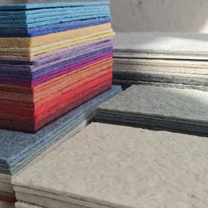 K&ouml;nnte beinhalten: Gestapelte Filzquadrate in verschiedenen Farben, von Blau &uuml;ber Gelb und Lila bis hin zu Orange und Rot. Die Quadrate sind in Schichten angeordnet und bilden einen Farbverlauf. Zus&auml;tzliche Stapel grauer und cremefarbener Filzquadrate sind ebenfalls zu sehen.
