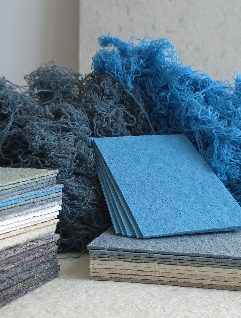 K&ouml;nnte beinhalten: Stapel quadratischer, strukturierter Filzstoff in Blau und Grau. Im Hintergrund befindet sich ein Haufen blauen Garns. Zus&auml;tzliche Stapel Filz in verschiedenen Farben sind ebenfalls sichtbar.