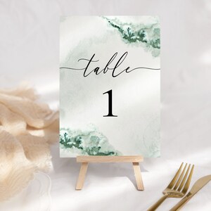 May include: A white table number card with a green watercolor design and the text "table 1" in black.