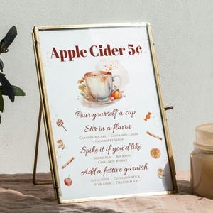 May include: A gold-framed sign with a watercolor illustration of a cup of apple cider. The sign reads "Apple Cider 5c" and includes instructions for adding flavors and garnishes.