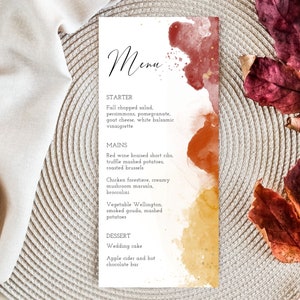 May include: A fall-themed menu with watercolor splashes in shades of red, orange, and yellow. The menu lists a starter, main courses, and dessert options. The text reads: "Menu, Starter, Fall chopped salad, persimmons, pomegranate, goat cheese, white balsamic vinaigrette, Mains, Red wine braised short ribs, truffle mashed potatoes, roasted brussels, Chicken forestiere, creamy mushroom marsala, broccolini, Vegetable Wellington, smoked gouda, mashed potatoes, Dessert, Wedding cake, Apple cider and hot chocolate bar."