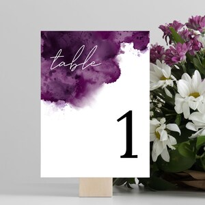 Puede incluir: Tarjeta de número de mesa de acuarela púrpura con la palabra "mesa" y el número "1" en texto blanco.