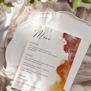 May include: A wedding menu card with a watercolor background in shades of red, orange, and yellow. The menu includes a starter, main courses, and dessert options. The text reads: "Menu, Starter, Fall chopped salad, persimmons, pomegranate, goat cheese, white balsamic vinaigrette, Mains, Red wine braised short ribs, truffle mashed potatoes, roasted brussels, Chicken forestiere, creamy mushroom marsala, broccolini, Vegetable Wellington, smoked gouda, mashed potatoes, Dessert, Wedding cake, Apple cider and hot chocolate bar."