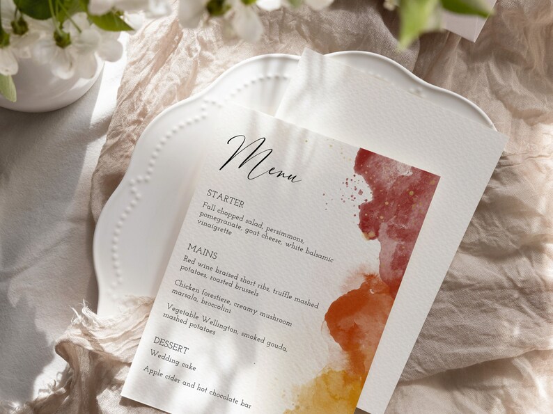May include: A wedding menu card with a watercolor background in shades of red, orange, and yellow. The menu includes a starter, main courses, and dessert options. The text reads: "Menu, Starter, Fall chopped salad, persimmons, pomegranate, goat cheese, white balsamic vinaigrette, Mains, Red wine braised short ribs, truffle mashed potatoes, roasted brussels, Chicken forestiere, creamy mushroom marsala, broccolini, Vegetable Wellington, smoked gouda, mashed potatoes, Dessert, Wedding cake, Apple cider and hot chocolate bar."