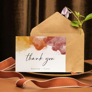 May include: A thank you card with a watercolor background in shades of yellow, orange, and red. The card says "thank you" in black script and "Kennedy - Curtis" in black print.