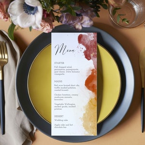 May include: A wedding menu card with a watercolor background in shades of red, orange, and yellow. The menu includes a starter, main courses, and dessert options. The text reads: "Menu", "Starter", "Fall chopped salad, persimmons, pomegranate, goat cheese, white balsamic vinaigrette", "Mains", "Red wine braised short ribs, truffle mashed potatoes, roasted brussels", "Chicken forestiere, creamy mushroom marsala, broccolini", "Vegetable Wellington, smoked gouda, mashed potatoes", "Dessert", "Wedding cake", "Apple cider and hot chocolate bar".