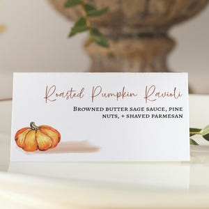 K&ouml;nnte beinhalten: Eine wei&szlig;e Karte mit dem Text "Roasted Pumpkin Ravioli" in brauner Schrift. Die Karte enth&auml;lt auch den Text "Browned Butter Sage Sauce, Pine Nuts, + Shaved Parmesan" in schwarzer Schrift. Eine Aquarell-Illustration eines K&uuml;rbisses befindet sich in der linken unteren Ecke der Karte.
