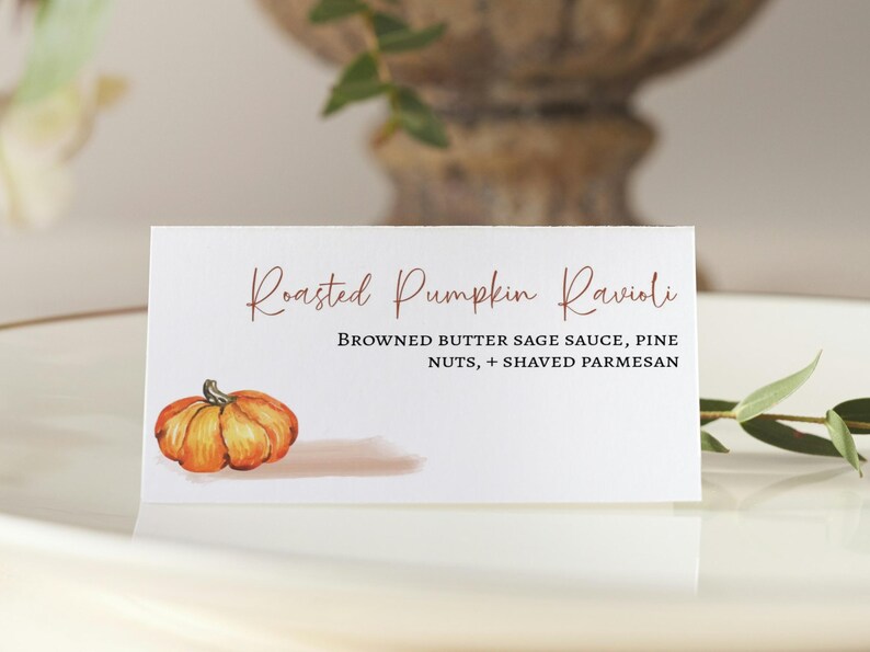 K&ouml;nnte beinhalten: Eine wei&szlig;e Karte mit dem Text "Roasted Pumpkin Ravioli" in brauner Schrift. Die Karte enth&auml;lt auch den Text "Browned Butter Sage Sauce, Pine Nuts, + Shaved Parmesan" in schwarzer Schrift. Eine Aquarell-Illustration eines K&uuml;rbisses befindet sich in der linken unteren Ecke der Karte.