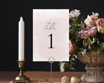 May include: A pink watercolor table number card with the text "Table 1" in black calligraphy. The card is on a metal stand and is surrounded by flowers.