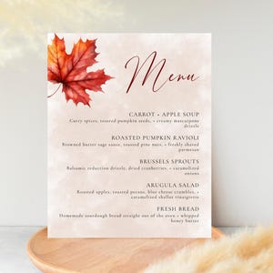 Könnte beinhalten: Eine Menükarte mit Aquarellhintergrund und einer Illustration eines roten und orangefarbenen Ahornblattes. Das Wort "Menü" ist in Schreibschrift geschrieben. Die Speisekarte listet Gerichte wie Karotten-Apfel-Suppe und geröstete Kürbis-Ravioli auf.