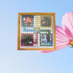 May include: Framed board game in a wooden frame. The game board has colourful squares and images of people. The text "Original Ludo Game" is visible. The background is a light blue sky with a pink flower.
