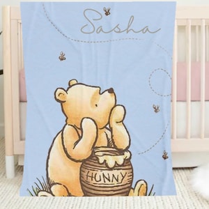 May include: A blue baby blanket with a Winnie the Pooh illustration. Pooh is sitting with a pot of honey that says "Hunny" on it. The blanket has the name "Sasha" written on it in a cursive font.