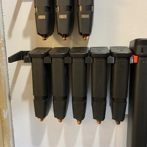 May include: A wall-mounted magazine holder with eight black magazine holders. The magazine holders are arranged in two rows, with four in each row. The top row is mounted on a blue plastic holder. The bottom row is mounted on a black plastic holder. Each magazine holder has a round, copper-colored bullet at the bottom.