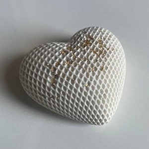 Op de afbeelding: Een wit hartvormig decoratief object met een textuur, roosterachtig oppervlak. Gouden vlokken zijn over de bovenkant verspreid. Het object is waarschijnlijk gemaakt van keramiek of een vergelijkbaar materiaal.