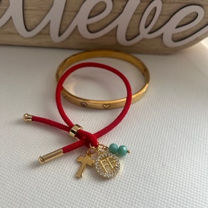 May include: A red string bracelet with a gold charm featuring a cross and a circle with Chinese characters. The bracelet also includes two small turquoise beads.
