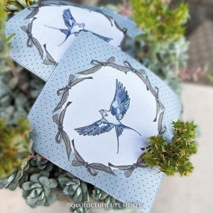 Könnte beinhalten: Zwei quadratische Stoffstücke mit hellblauem gepunkteten Rand und einem weißen Zentrum mit einem blau-weißen Vogel im Flug. Der Rand hat ein graues Schleifendesign. Der Text "acufactum UTE MENZE" befindet sich unten.