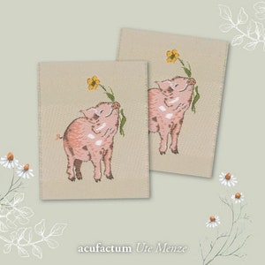 Könnte beinhalten: Zwei beige Stoffpatches mit gestickten rosa Ferkeln, die gelbe Blumen halten. Der Text "acufractum Ute Menze" ist unter den Patches gedruckt.