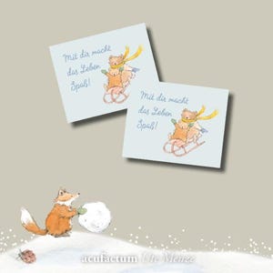 Könnte beinhalten: Zwei hellblaue Karten mit einer Aquarellillustration von Bären auf einem Schlitten und dem Text "Mit dir macht das Leben Spaß!". Ein Fuchs rollt im Vordergrund einen Schneeball. Der Name des Künstlers steht unten.